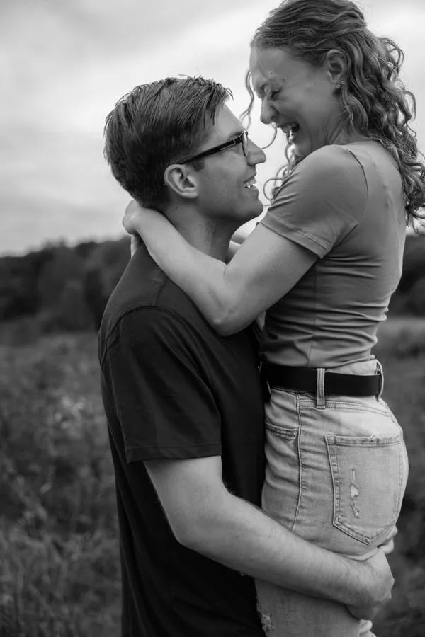 Man in t-shirt and glasses lifting up curly-haired girl in jeans laughing in black and white